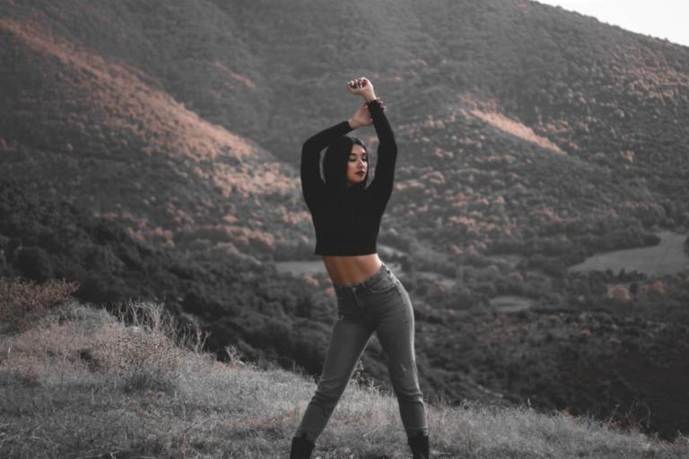A confident young woman in a black crop top and jeans stands on a grassy mountain peak with arms raised, embodying strength and vitality against a backdrop of vast, forested hills.