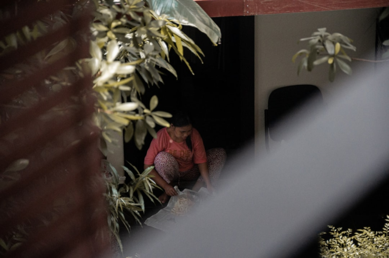 A woman in a pink shirt crouches among lush green plants in a shadowed doorway, intently sorting items from a bag, embodying a focused calm conducive to deeper meditation and mental clarity.