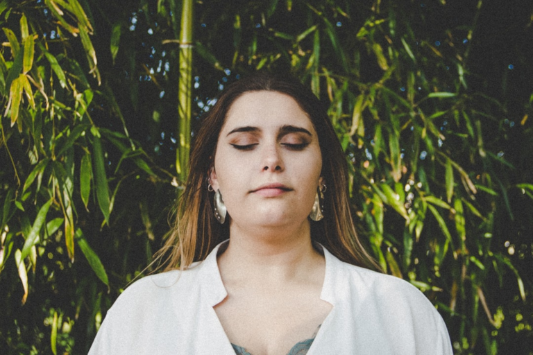 A woman with closed eyes and a peaceful expression stands against a lush green bamboo backdrop, conveying a moment of ocular relaxation.
