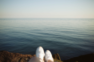 Relaxed feet on a rocky shore overlook a tranquil blue ocean under a vast, clear sky, symbolizing the peace and rest crucial for enhanced brain performance.