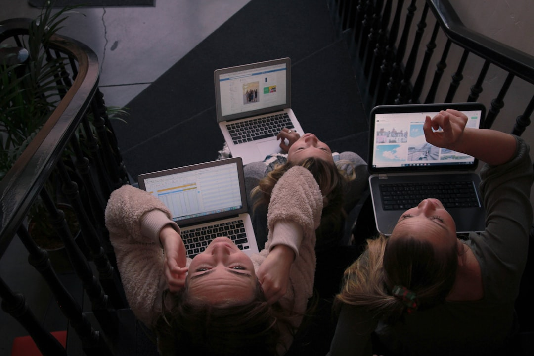 Three young individuals lie casually on a dark floor, engrossed in their bright laptop screens next to a staircase, reflecting typical extended device use and the need for optimized screen settings for eye health.