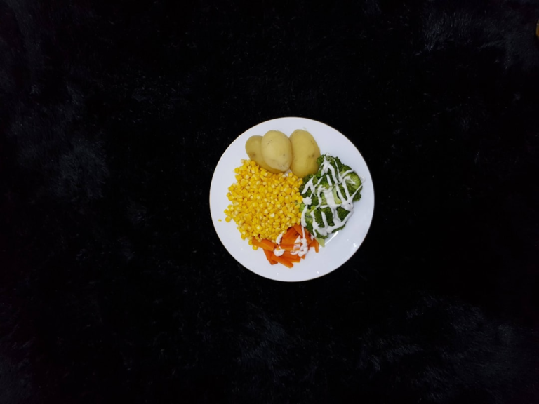 Balanced plate of steamed broccoli with a creamy drizzle, corn, carrots, and potatoes set against a dark, textured surface, embodying a healthy, fiber-rich diet for optimal gut microbiome.