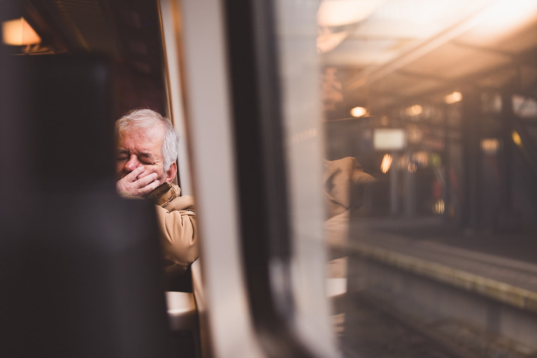 An older man, appearing weary, covers his mouth while resting his eyes on public transport, with a blurred train station visible through the window.