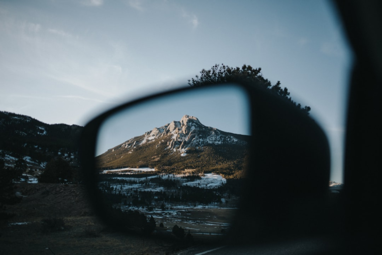 A prominent snow-capped mountain and forested landscape are sharply reflected in a car's side mirror under a clear sky, representing a tranquil view and the importance of clear vision.
