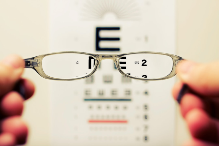 Hands hold transparent prescription eyeglasses, bringing a blurry eye chart into sharp focus through the lenses, illustrating improved vision and eye health.