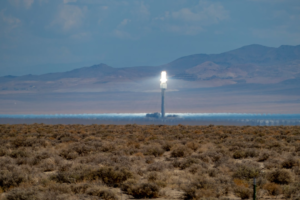 A brilliant solar power tower shines intensely, its concentrated light illuminating a vast desert expanse with distant mountains under a partly cloudy sky, embodying the harnessing of light for energy.