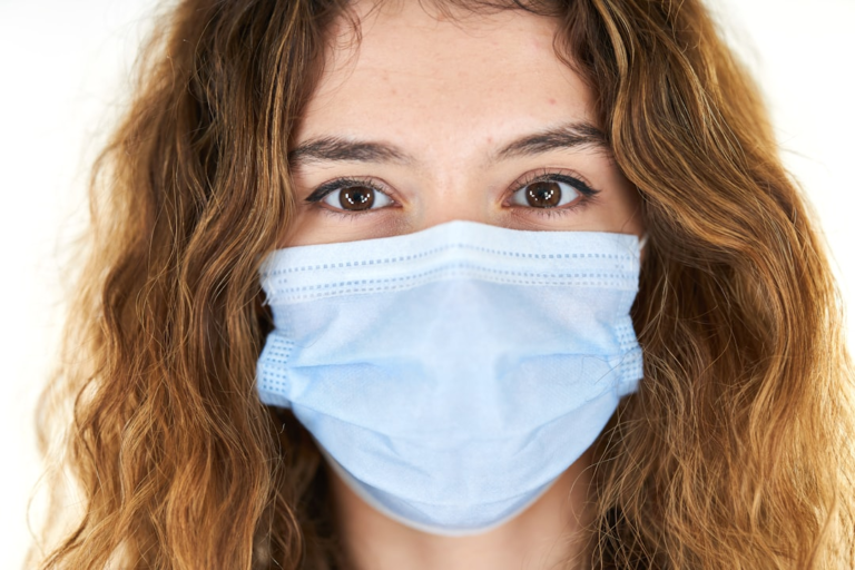 A young woman with attentive brown eyes and wavy brown hair wears a blue medical mask, representing focus on vision health and advanced eye care.