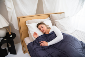 Serene woman sleeps soundly on her side in a light-filled modern bedroom, comfortably covered by a dark blue weighted blanket on a natural wood bed, representing deep rest for metabolic harmony.