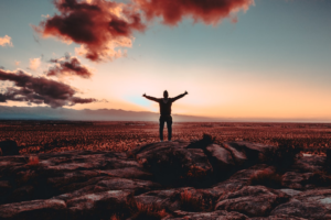 Silhouette of a person with outstretched arms standing triumphantly on a rocky summit, overlooking a vast desert landscape under a vibrant sunset sky.