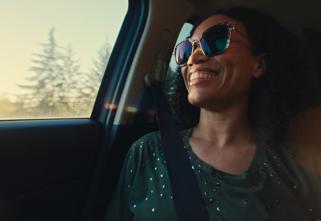 A smiling woman wearing stylish blue light blocking glasses looks contentedly out a car window at a blurred, sunlit natural landscape.