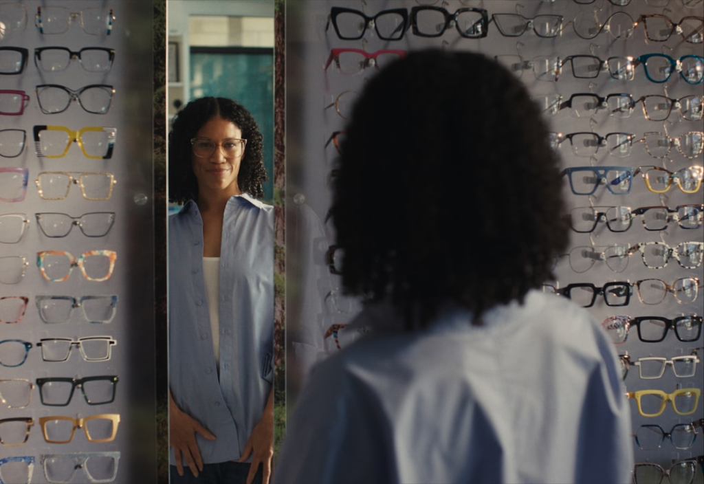 A smiling woman wearing stylish glasses looks at her reflection in a mirror, surrounded by rows of diverse frames in an optical store, highlighting choices for blue light blocking eyewear.