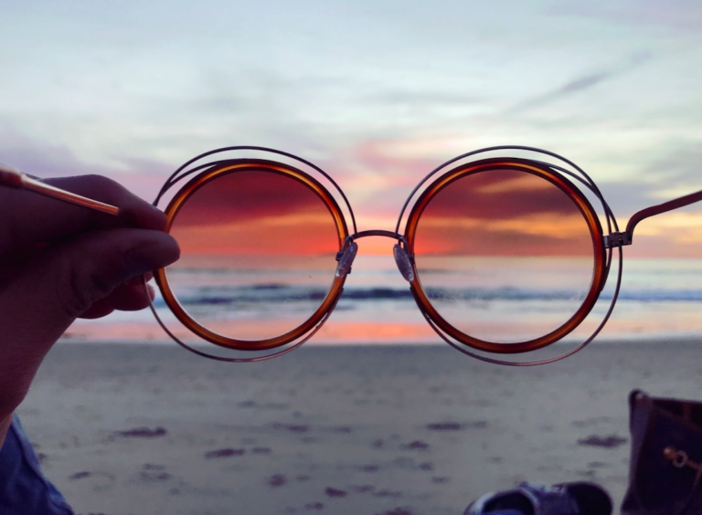 Hand holding round, vintage-style eyeglasses, framing a vibrant orange and purple sunset over a tranquil ocean beach, suggesting a fresh perspective on natural vision.