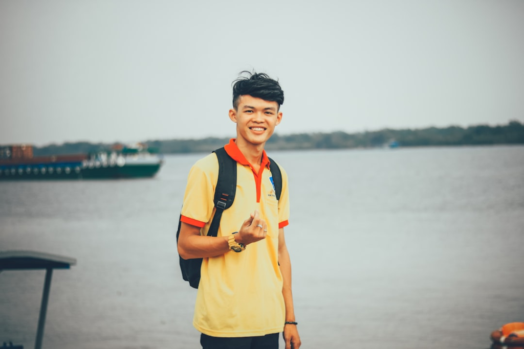 Vibrant young man with a cheerful smile and backpack stands by a wide river with a distant cargo ship, reflecting the vitality of a lifestyle optimized for longevity.