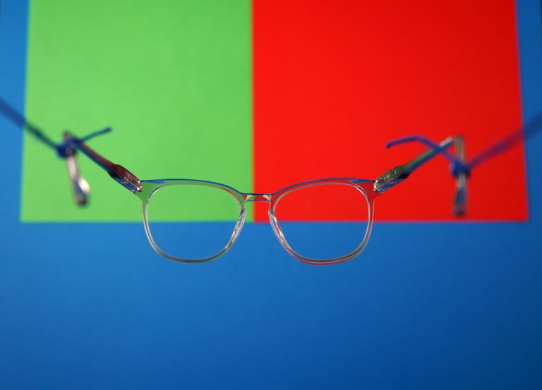 Clear-framed eyeglasses are suspended against a vibrant, color-blocked background of red and green squares above a blue surface, symbolizing visual clarity and optimized brain-eye connection.