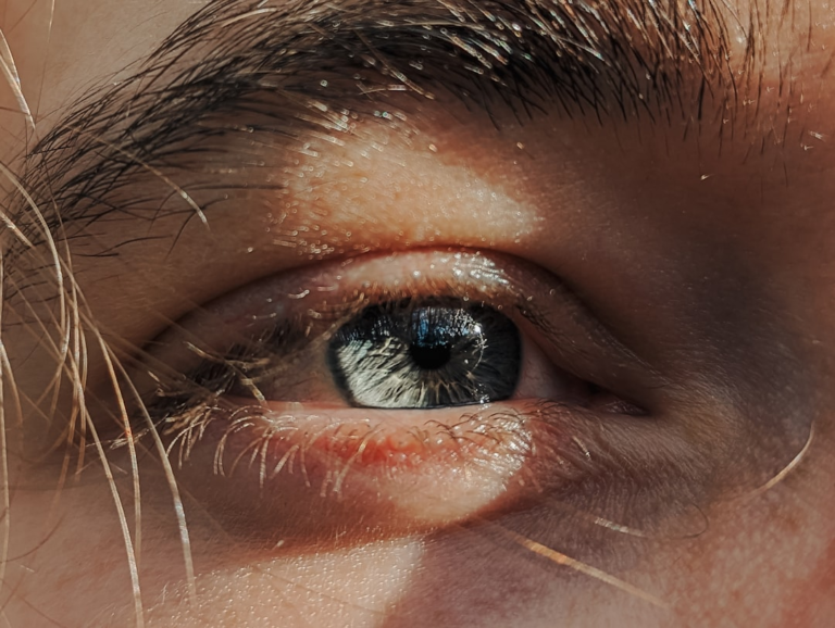 Detailed close-up of a human eye with a blue-grey iris and prominent eyelashes, illuminated by a strong light, visually representing ocular strain and the connection between stress and vision.