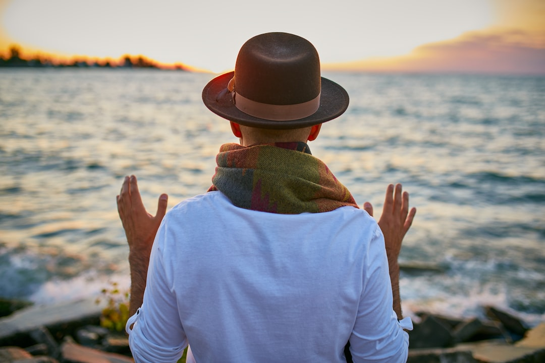 Person in a fedora and colorful scarf stands before a tranquil sunset over calm waters, hands open in a reflective pose, symbolizing peace and the restorative rhythms of nature.
