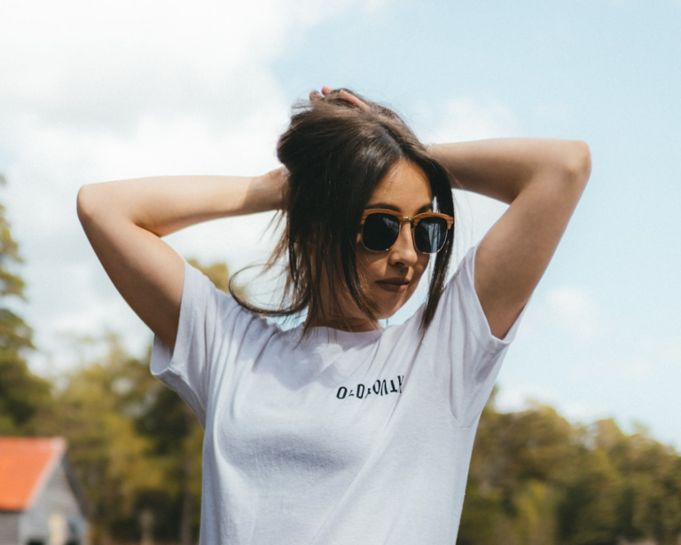 Young woman wearing trendy sunglasses, hands in her hair, stands outdoors against a bright sky, emphasizing the necessity of protecting eyes from sun damage.