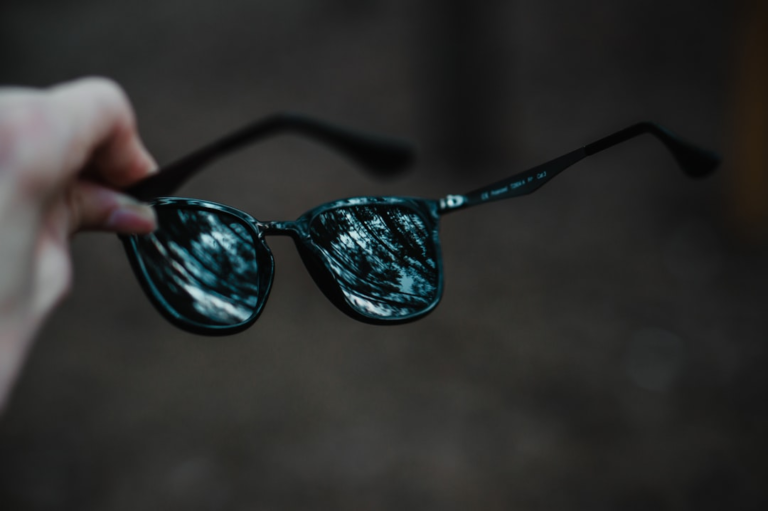 A hand holds dark sunglasses whose lenses brightly reflect a vibrant, natural landscape, symbolizing the promise of clearer vision from emerging AMD treatments.
