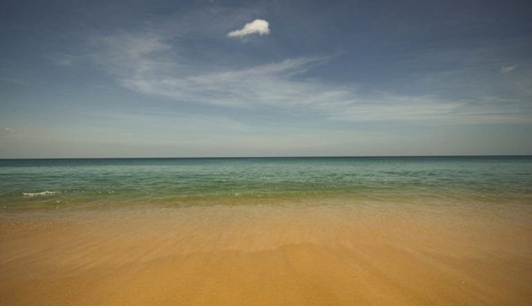Golden sandy beach gently meets serene turquoise ocean waves under a vast blue sky with a soft white cloud, creating a scene of profound tranquility.