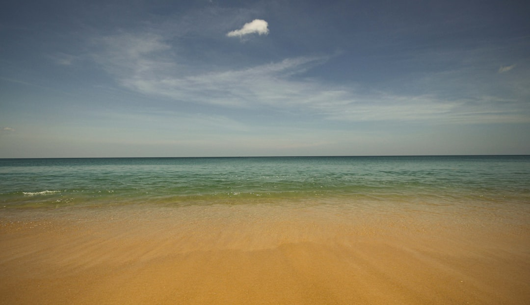 Golden sandy beach gently meets serene turquoise ocean waves under a vast blue sky with a soft white cloud, creating a scene of profound tranquility.