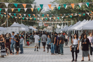 A diverse crowd browses a vibrant outdoor market lined with white tents offering healthy and natural products, adorned with festive bunting, embodying a community focused on holistic wellness.