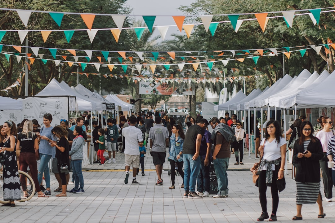 A diverse crowd browses a vibrant outdoor market lined with white tents offering healthy and natural products, adorned with festive bunting, embodying a community focused on holistic wellness.