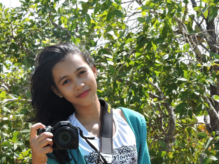 A smiling young woman with dark hair holds a Kodak camera, standing joyfully amidst lush green foliage, embodying an active lifestyle essential for extended healthspan.