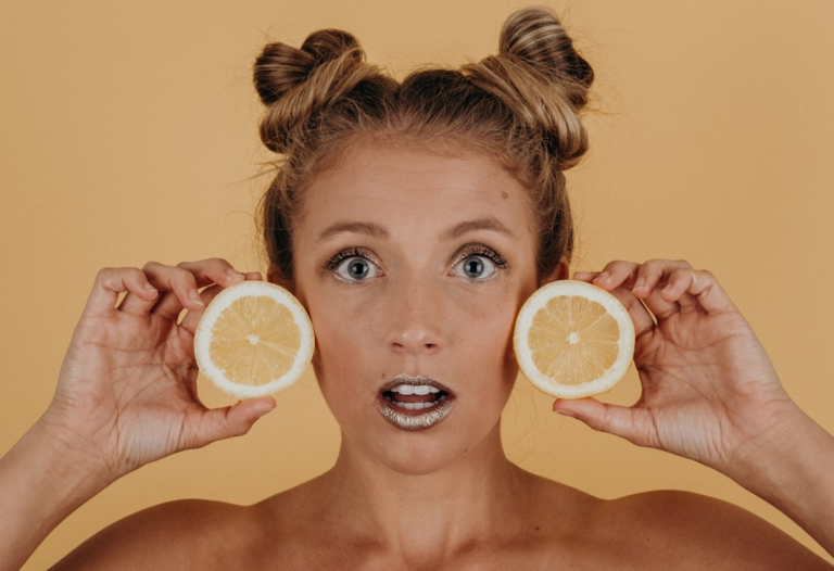 Surprised woman with sparkling lips and space buns holds two vibrant yellow lemon halves near her face, symbolizing fresh nutrition and ingredients beneficial for healthy eyes and macular health.