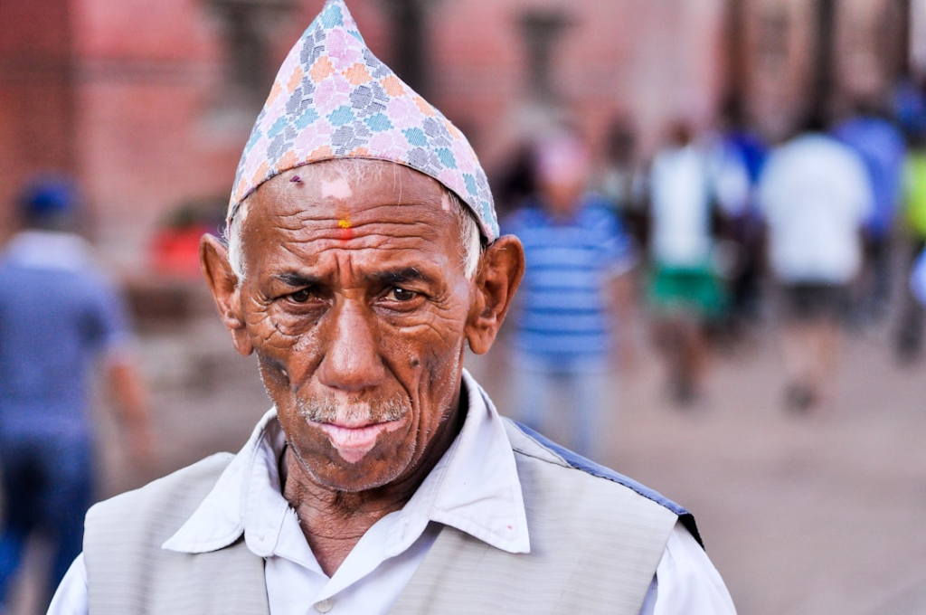 An elderly man with a deeply wrinkled face, a patterned hat, and a red bindi gazes intently, his features embodying the journey and wisdom of longevity against a blurred urban background.