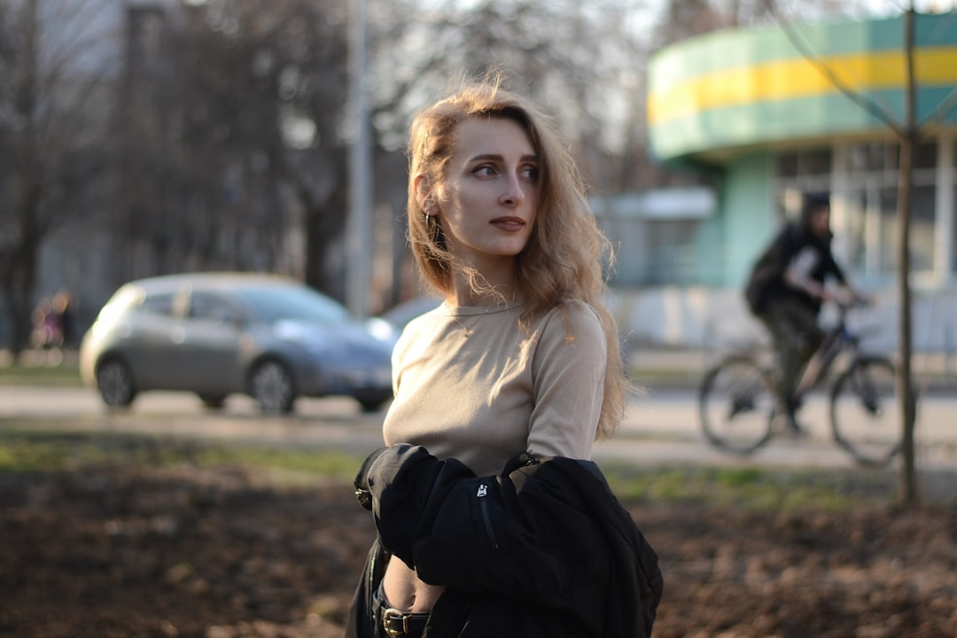 A young woman with a radiant complexion and long, wavy blonde hair looks thoughtfully over her shoulder in a sunlit urban setting.