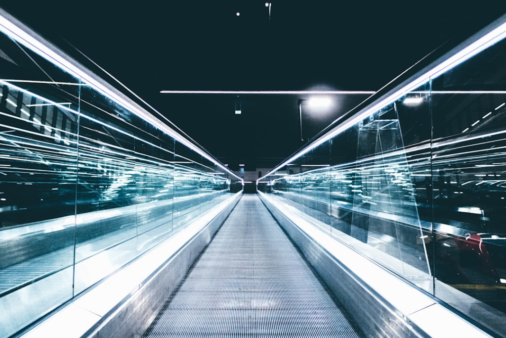 An illuminated moving walkway stretches into a dark distance, flanked by reflective glass walls adorned with luminescent streaks of blue and white light, symbolizing a transformative journey for programming your reality.