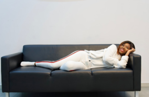 A contented young woman in comfortable white clothing relaxes on a black sofa, embodying the revitalizing effects of rest and healthy sleep.