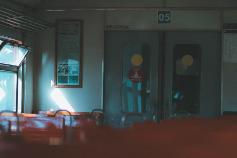 Quiet train car interior with rows of empty red seats, a bright sunbeam illuminating the wall from an open window, and a closed grey door displaying Russian signage.