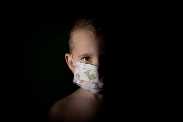 Young child's eye, clearly visible from behind a dinosaur-patterned face mask in dramatic shadow, drawing attention to eye health and vision.