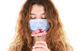 A serene young woman with closed eyes and long wavy hair, wearing a medical mask, gently holds a pink flower, representing a peaceful moment of self-care and eye relaxation.