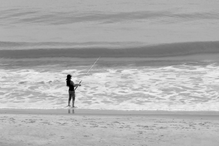 A solitary angler stands focused in the foamy ocean surf, casting a line into the rolling waves on a tranquil black and white beach, reflecting the patient and persistent pursuit of natural energy.
