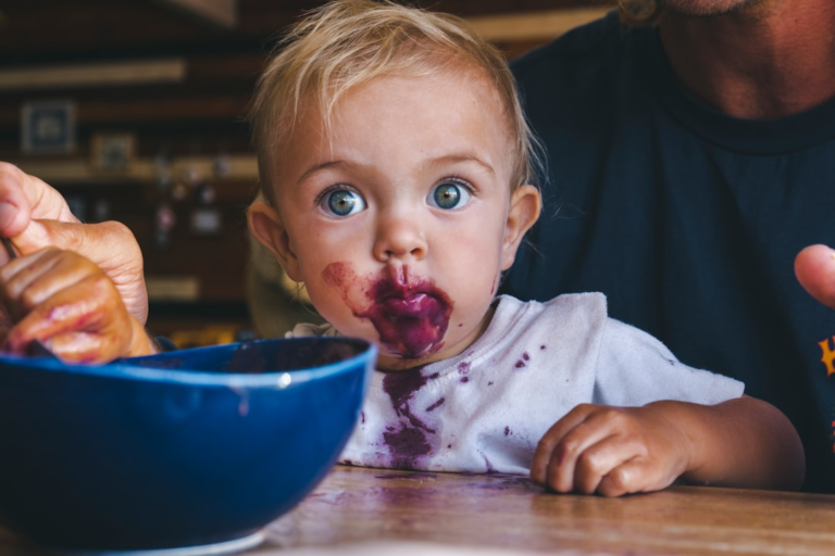 A blonde baby with striking bright blue eyes and a face smudged with purple berry food gazes curiously at the camera, highlighting the role of diet in developing healthy vision.