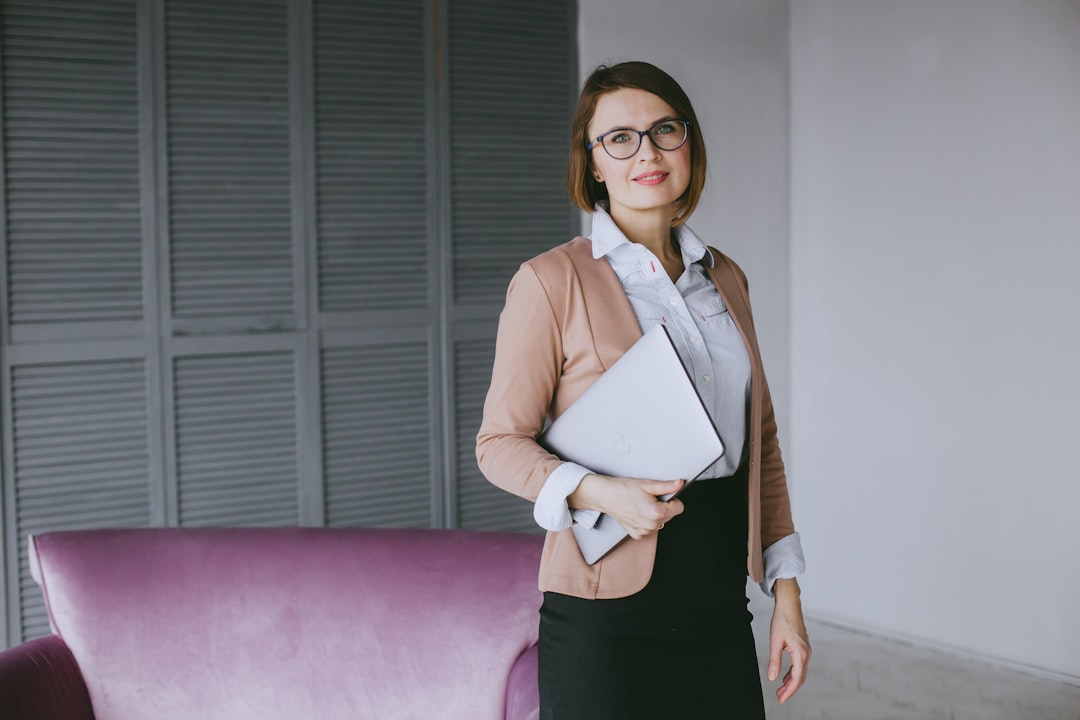 A smiling professional woman wearing blue light glasses and a beige blazer holds a silver laptop in a bright modern office setting.