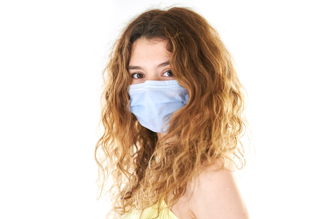 A young woman with prominent brown eyes and flowing auburn hair, wearing a light blue face mask, looks directly forward on a stark white background, emphasizing eye health.